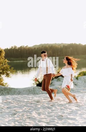 Couple heureux courant sur le sable, coucher de soleil, couple élégant avec une valise et des fleurs dans un panier. Banque D'Images