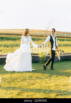 Promenade romantique de jeunes mariés amoureux dans le champ tenant la main l'un de l'autre au coucher du soleil Banque D'Images