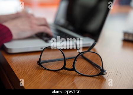 Lunettes à monture noire sur un bureau en bois, vue rapprochée. Blur les mains des femmes sur un ordinateur portable clavier, Banque D'Images