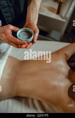 Un bol avec de l'huile chaude infusée aux herbes pour le massage du dos dans les mains d'un thérapeute masculin Banque D'Images