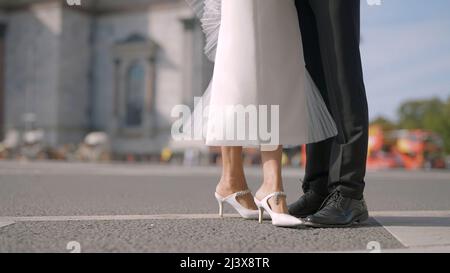 Jambes de mariée et marié élégant sur le fond de la ville rue d'été avec des voitures de conduite. Action. Homme en costume noir et femme en robe blanche. Banque D'Images