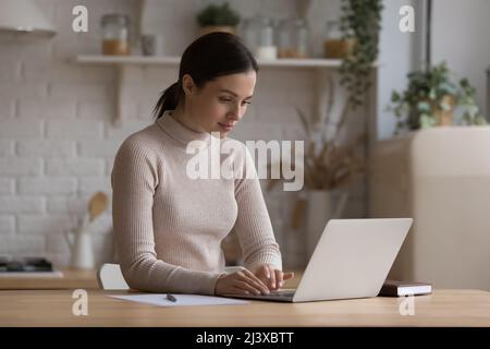 Jolie jeune fille freelance millénaire utilisant un ordinateur portable dans la cuisine à la maison Banque D'Images