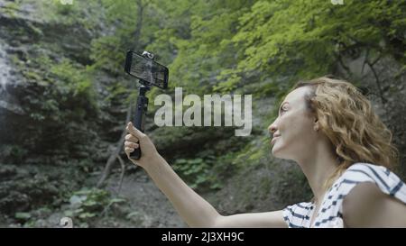 Femme prenant des photos ou filant des montagnes et des chutes d'eau sur son smartphone à l'aide d'un trépied. Action. Femme voyageur avec un bâton de selfie et un Banque D'Images