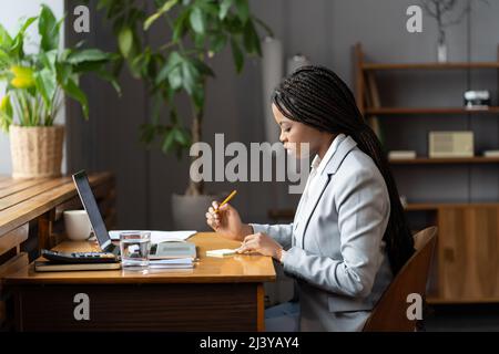 Assistante de bureau africaine ciblée écrivant quelque chose sur le post-it, planifiant la journée de travail Banque D'Images