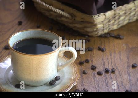 Une tasse de café noir sur une soucoupe à rebord marron avec 3 grains de café. Les grains de café renversés se trouvent sur le plan d'examen texturé brun à côté d'un plateau en osier Banque D'Images