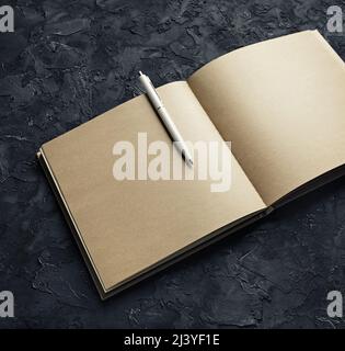 Photo d'un livre kraft ouvert avec des pages vierges et un stylo sur fond de plâtre noir. Banque D'Images