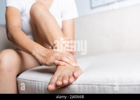 Douleur au pied, femme asiatique assise sur un canapé tenir sa blessure à la cheville sensation de douleur dans son pied à la maison, femme souffrant de douleur aux pieds utiliser massage des mains se détendre Banque D'Images