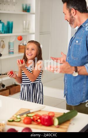 Un dîner et un spectacle maintenant c'est bon pour votre argent. Photo d'une adorable petite fille qui jongle avec des tomates tout en cuisinant avec son père à la maison. Banque D'Images