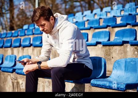 Homme dans les vêtements de sport assis sur le siège du stade et de navigation smartphone, Homme sportif vérifier la montre de fitness après les exercices de sport Banque D'Images