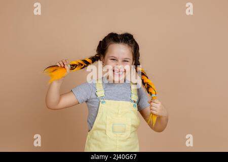 Jeune fille rayonnante et satisfaite ayant des tresses kanekalon de jaune, riant et tirant des queues de porc regardant la caméra souriant portant une combinaison jaune Banque D'Images