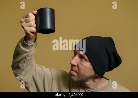 Un jeune homme drôle regarde avec surprise dans une tasse noire inversée. Portrait d'un européen en casquette noire Banque D'Images