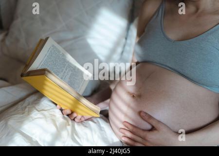 Une femme enceinte lit un livre et se couche le ventre avec un bébé avant d'accoucher tout en se relaxant dans une chambre ensoleillée et lumineuse, couchée sur un lit. Mise au point sélectionnée. La santé des femmes, la grossesse, la conception, l'accouchement concept. Photo de haute qualité Banque D'Images