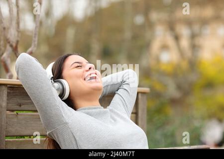 Une femme heureuse qui écoute de la musique avec un casque se détend sur un banc dans un parc Banque D'Images