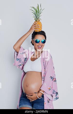 Shes une belle-mère de pomme à être. Photo studio d'une belle jeune femme enceinte tenant un ananas sur sa tête sur un fond gris. Banque D'Images