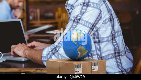 Globe au-dessus de la boîte de livraison contre la section médiane d'une personne utilisant un ordinateur portable dans un café Banque D'Images