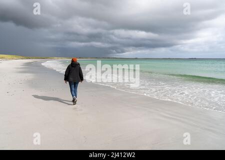 Une femme marche le long d'une plage de sable blanc déserte, adossée à des machair, avec des nuages de tempête à l'horizon. Banque D'Images