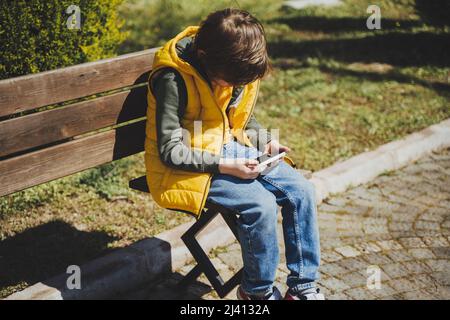 Un écolier joue au jeu de son téléphone portable assis sur un banc dans le parc de la ville. Garçon enfant dans la veste jaune et le sweat à capuche vert utilise le gadget mobile pour discuter avec des amis Banque D'Images