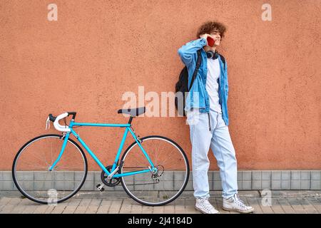 Jeune homme souriant aux cheveux bouclés avec un casque debout au mur à l'extérieur, parlant sur un téléphone portable à côté de son vélo bleu. Banque D'Images