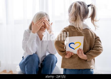 fille avec queue de cheval tenant une carte avec coeur bleu et jaune près de maman heureuse obscurcissant le visage avec les mains Banque D'Images