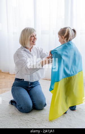 femme blonde souriante couvrant petite fille avec drapeau ukrainien à la maison Banque D'Images