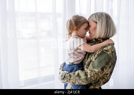 vue latérale de la petite fille embrassant la mère en uniforme militaire à la maison Banque D'Images