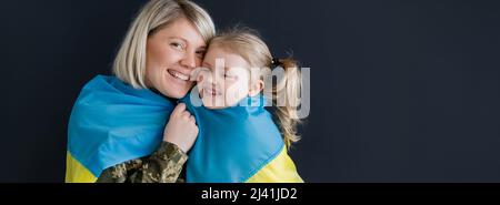 bonne femme regardant l'appareil photo près de la fille et drapeau ukrainien isolé sur noir, bannière Banque D'Images