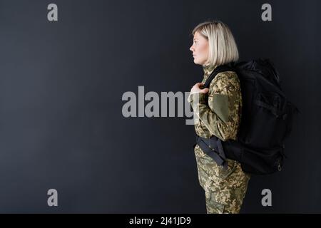 vue latérale d'une femme militaire blonde avec sac à dos isolé sur le noir Banque D'Images