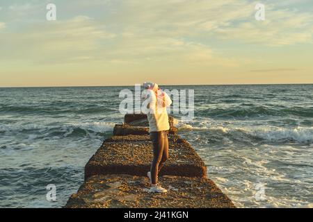 Une femme aux cheveux blonds dans un chapeau et un manteau de fourrure marche le long de la mer le long d'un brise-lames en pierre Banque D'Images