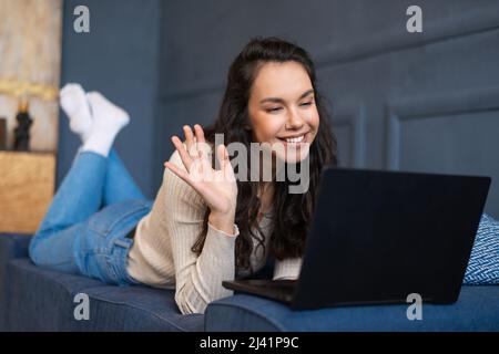 Une femme enthousiaste visiophonie via un ordinateur portable, se coucou à la webcam de l'ordinateur, couchée sur un canapé dans le salon Banque D'Images