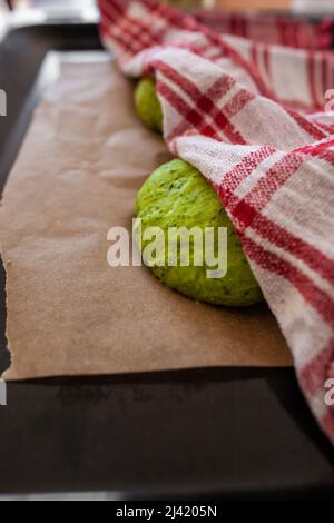 Pâte verte sous une serviette de cuisine à carreaux sur du papier de cuisson brun sur une plaque de cuisson noire pâte à pain à pâte levée recouverte d'un chiffon de cuisine blanc rouge Banque D'Images