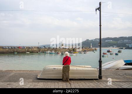 Une femme âgée aux cheveux blancs et au pelage rouge vif se trouve devant un bateau retourné, face à Smeaton's Pier à St Ives, Cornwall, Royaume-Uni. Banque D'Images