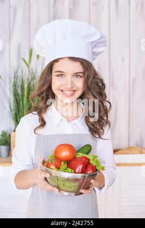 Jeune fille cuisant dans la cuisine avec des légumes. Joli portrait d'enfant. Étudiant chef. Une jolie brunette. L'émotion des enfants. Les gens qui cuisinent dans un tablier de cuisson. Fermer le menu de recette Banque D'Images