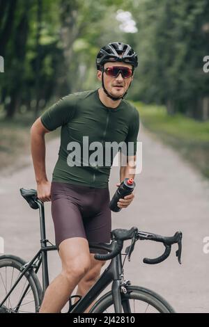 Beau jeune homme en vêtements d'activité, casque de sécurité et lunettes à miroir reposant sur le vélo après une promenade matinale au parc de la ville. Concept de personnes, passe-temps actif Banque D'Images