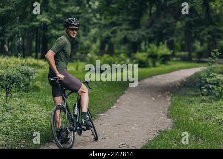 Homme fort assis sur le vélo et souriant sur l'appareil photo tout en prenant une pause après un entraînement actif sur l'air frais. Cycliste masculin portant des vêtements de sport, casque de sécurité Banque D'Images