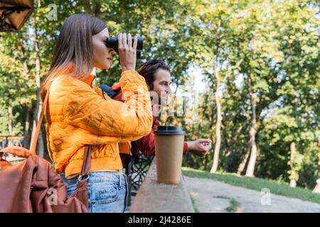 Jeune touriste regardant à travers des jumelles près d'un petit ami souriant et une tasse de papier à l'extérieur Banque D'Images