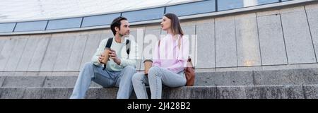 Les jeunes voyageurs avec un café à emporter et des sacs à dos qui parlent dans les escaliers près du bâtiment dans la rue urbaine, bannière Banque D'Images