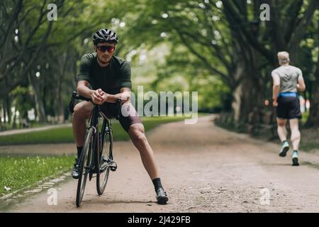 Homme caucasien avec des jambes musculaires assis sur le vélo de sport noir tout en prenant une pause de vélo à l'extérieur. Homme actif portant des vêtements de sport, un casque de sécurité Banque D'Images