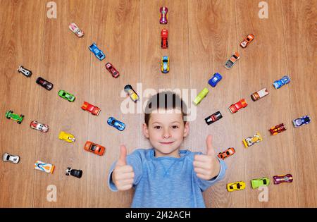 petit enfant un garçon dans un chandail bleu est souriant couché sur le plancher de bois sur le fond de beaucoup de voitures jouets Banque D'Images