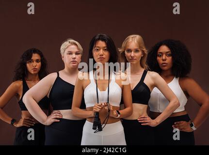 Cinq femmes en vêtements de sport regardant l'appareil photo. Les femelles avec différents types de corps et ethnies debout ensemble sur fond brun. Banque D'Images