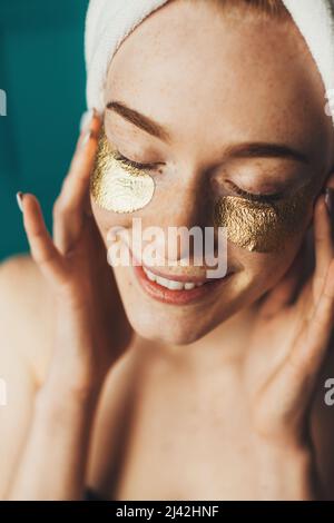 Après la douche, femme à tête rousse appliquant des taches dorées sous les yeux, touchant le visage avec les mains, posant à l'intérieur. Bonne humeur. Soins de la peau. Soins des yeux. Banque D'Images