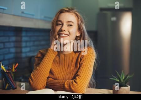 Femme à tête rouge assise à table avec un carnet et regardant l'appareil photo de rêve. Réfléchir à des idées créatives pour la recherche. Banque D'Images