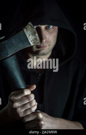 Portrait d'un jeune homme à capuchon avec des yeux bleus qui avec une hache dans sa main a un aspect inquiétant - foyer sur la lame Banque D'Images