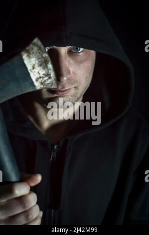 Portrait d'un jeune homme à capuchon avec des yeux bleus qui avec une hache dans sa main a un regard inquiétant - se concentrer sur l'oeil Banque D'Images