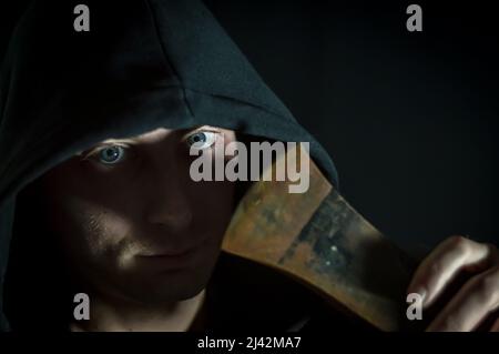 Portrait d'un jeune homme à capuchon avec des yeux bleus qui avec une hache dans sa main a un regard inquiétant - se concentrer sur les yeux Banque D'Images