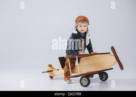 Un garçon heureux dans un casque et une veste de pilote se tient près d'un avion en bois. Portrait d'un pilote d'enfant, d'un enfant dans une veste en cuir. Jouets en bois. Plan ECO Banque D'Images