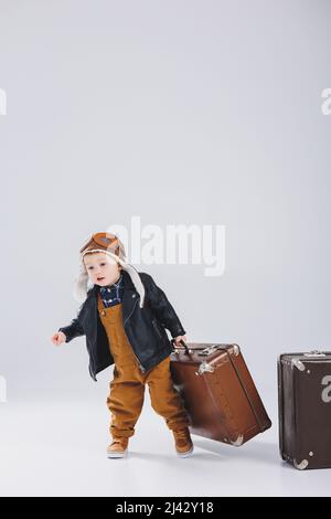 Un garçon heureux dans un casque et une veste de pilote portent une valise marron. Portrait d'un pilote d'enfant, d'un enfant dans une veste en cuir. Petit voyageur avec un Banque D'Images