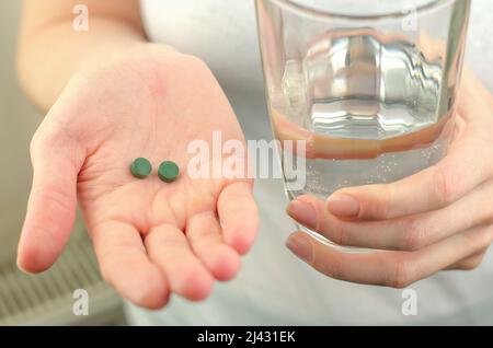 Deux pilules vertes de spiruline et un verre d'eau dans les mains des femmes, gros plan Banque D'Images