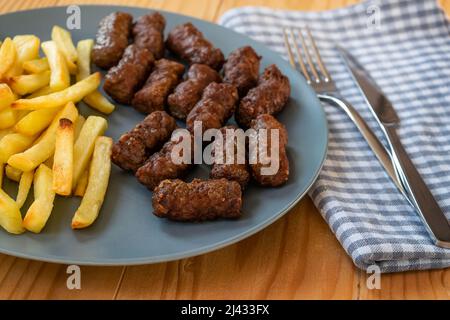 CEVAPi, plat traditionnel de viande hachée des balkans, servi avec des frites dans une assiette bleu marine sur une table en bois Banque D'Images