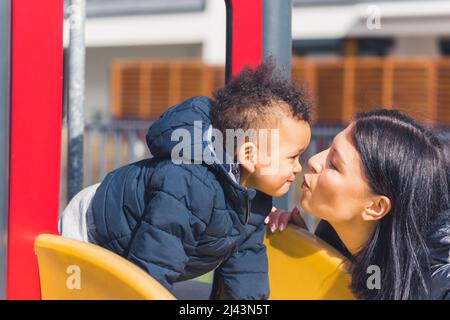 une femme aux cheveux foncés et un petit garçon mignon avec des cheveux bouclés noirs qui s'embrassent les uns les autres sur un cliché de diapositive de taille moyenne en extérieur. Photo de haute qualité Banque D'Images