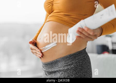 Soins de la peau pendant la grossesse. Femme enceinte appliquant de la crème sur l'estomac pour les marques d'étirement. Masque hydratant gros plan des mains et bouteille de lotion à la maison Banque D'Images